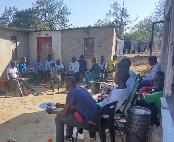 Group of men and boys seated in a circle in an outdoor courtyard, listening and talking during a community mental-health discussion.