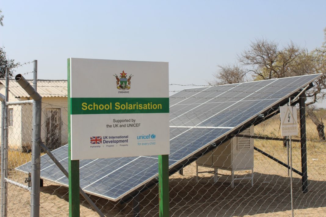 Ground-mounted solar panels behind a fence with a sign reading “School Solarisation — Supported by the UK and UNICEF” at a rural Zimbabwe school.