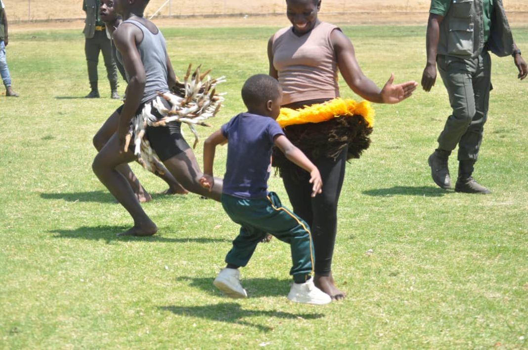 A young boy in a navy T-shirt and green track pants dances on a grass field with traditional dancers wearing feathered and tasseled costumes at a ZPCS event.