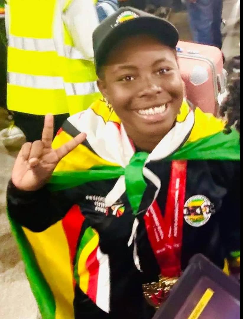 Smiling teenage karate athlete Anotida Hove draped in a Zimbabwean flag, wearing her team tracksuit and medal ribbons, holding up a peace sign at the airport.