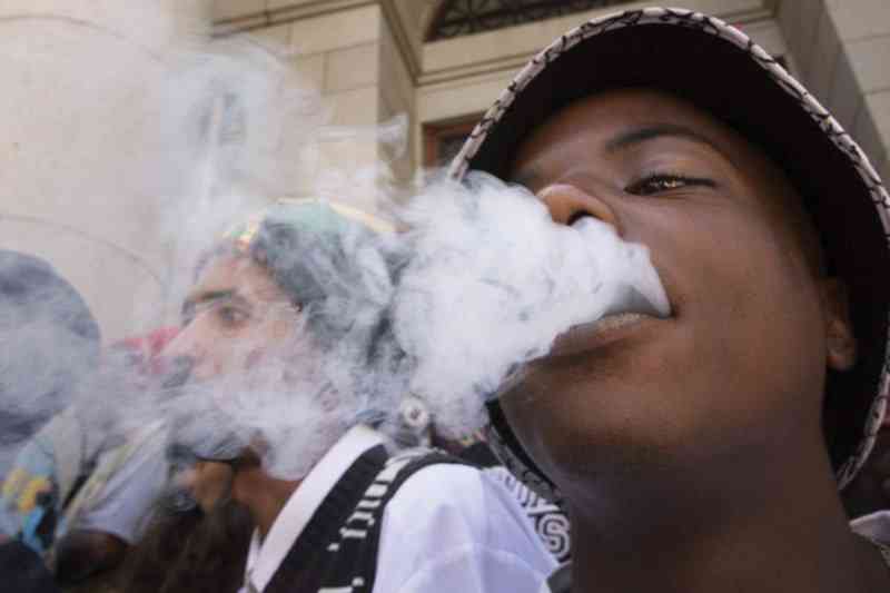Close-up of a teenage boy in a bucket hat exhaling thick smoke, with another young person blurred in the background.