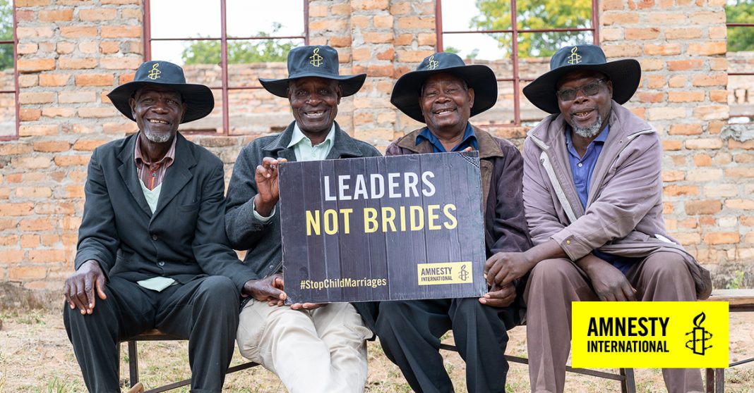 Four elderly men wearing hats sit on a bench holding a sign reading “Leaders Not Brides” during an Amnesty International campaign against child marriage, symbolising community support for ending forced marriages of girls.