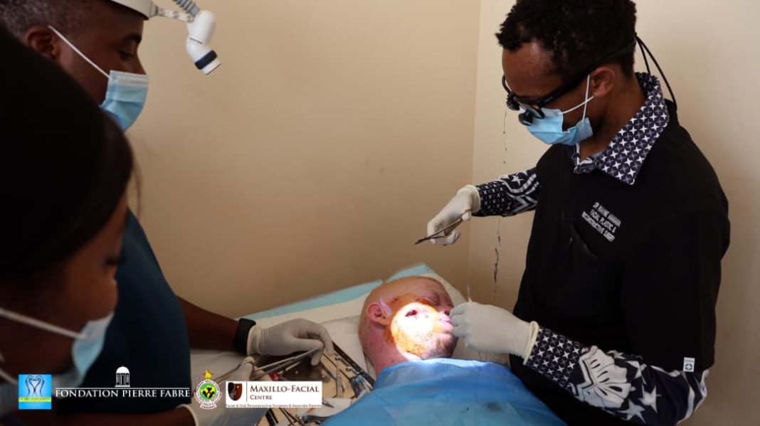 A masked clinician wearing gloves uses dental instruments and a bright examination light to work on a patient lying on a treatment bed, while two other masked staff members assist nearby.