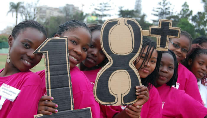 A group of school-aged girls wearing pink shirts stand together holding large cardboard signs reading “18+,” symbolising the legal age of marriage and advocacy against child marriage.