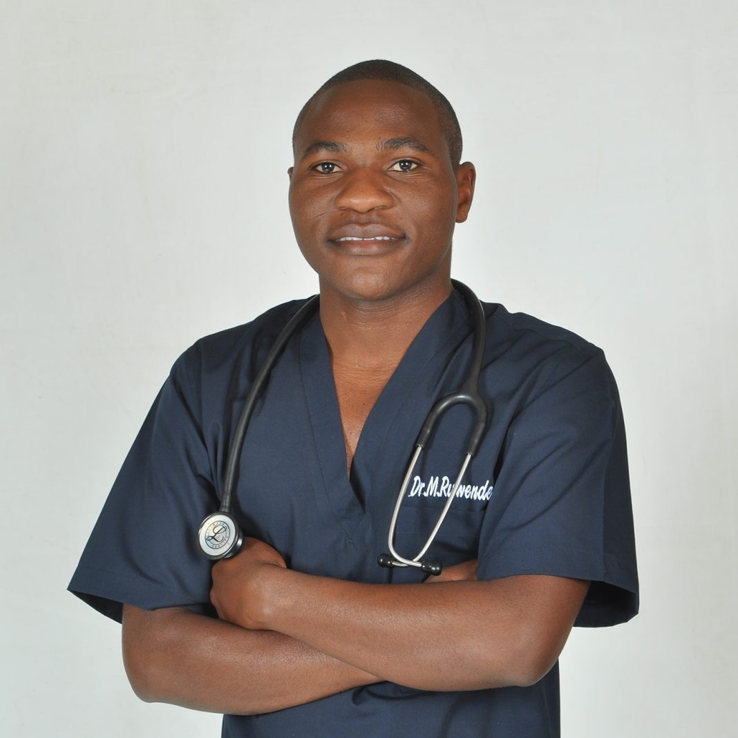Dr Misheck Ruwende, a Zimbabwean medical doctor, stands with arms crossed in navy scrubs with a stethoscope around his neck.
