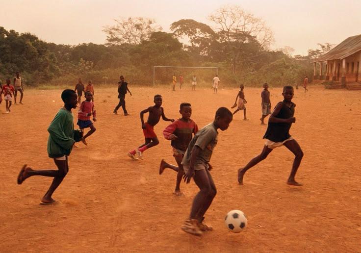Children playing football on a dusty field without formal sporting facilities.