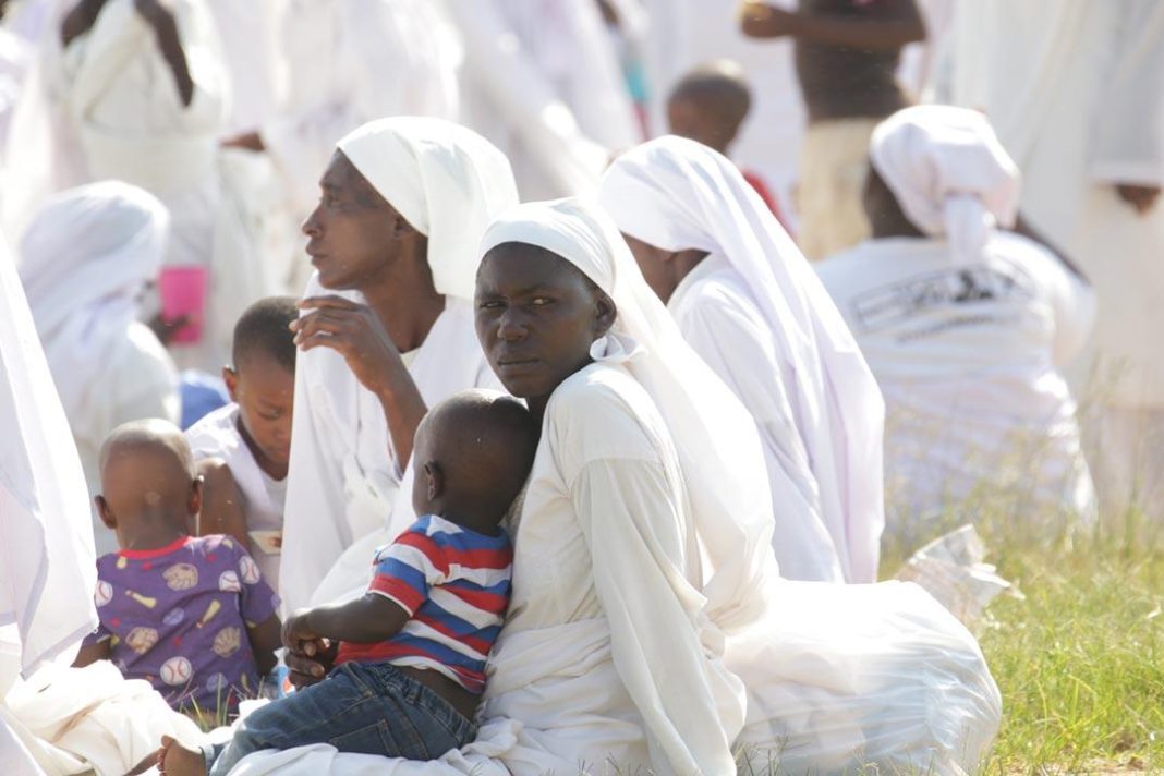 Apostolic church women dressed in white robes and headscarves sit with young children at an outdoor religious gathering.